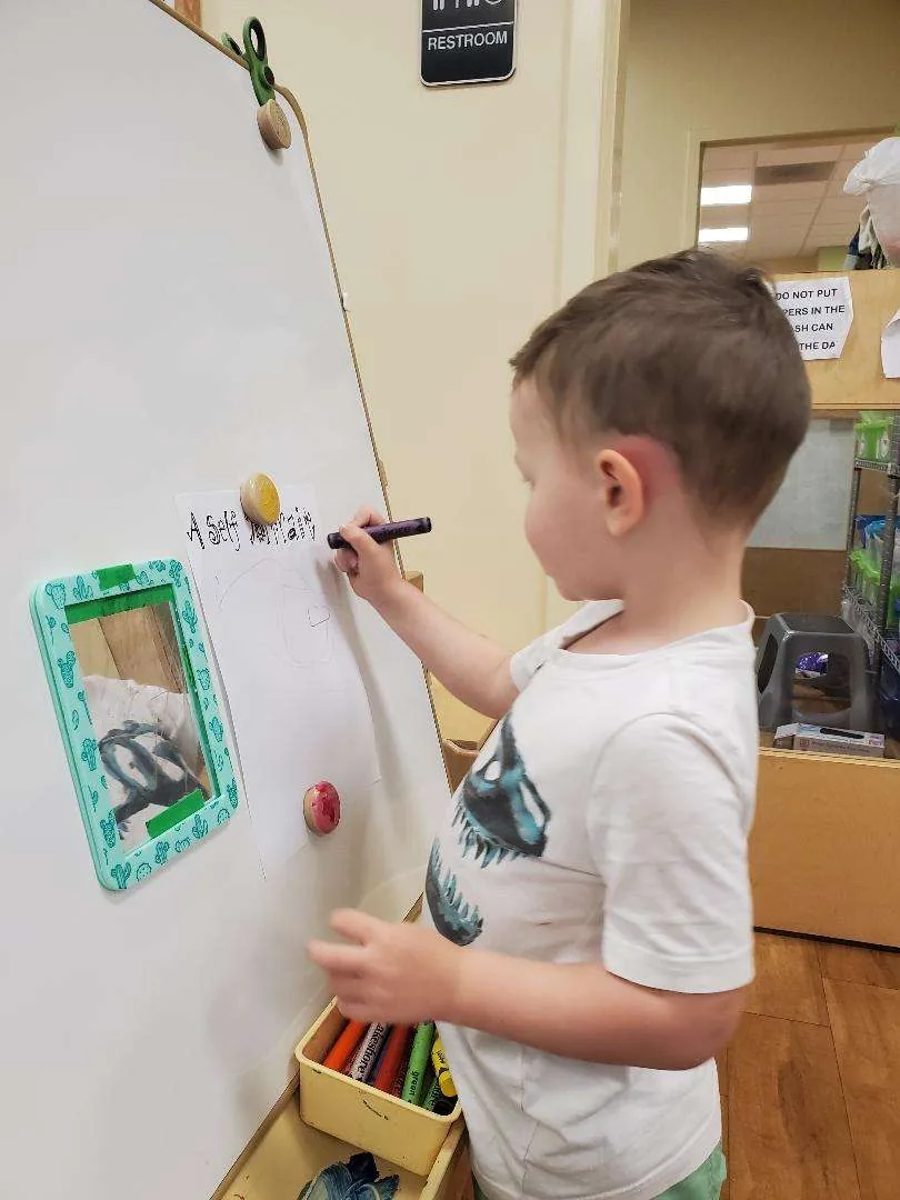 A preschooler drawing a self-portrait at the easel while looking in a hand mirror at Blossom Station in Acton, MA