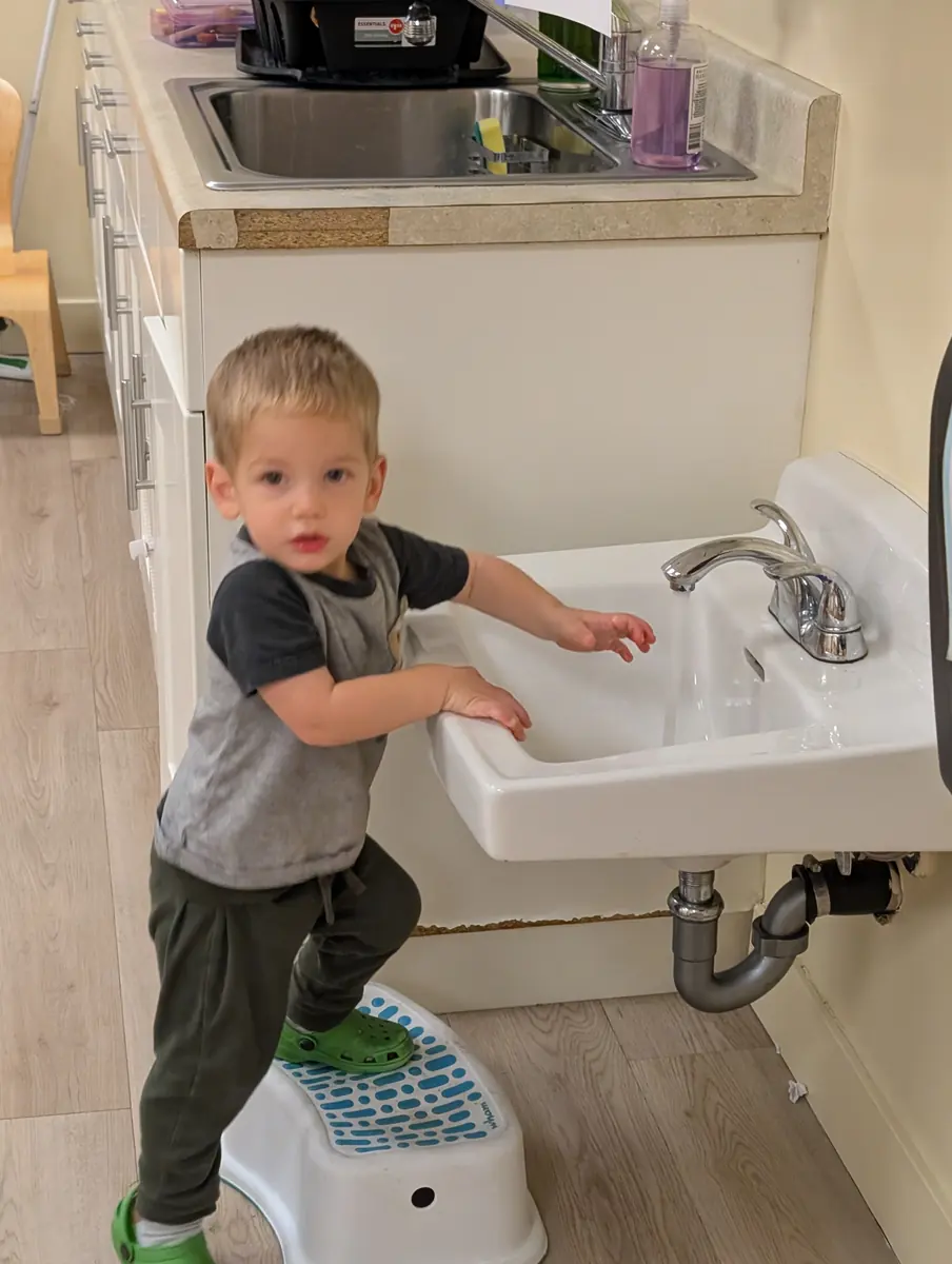 A toddler washing hands at a low sink, building self-help skills at Blossom Station in Acton, MA