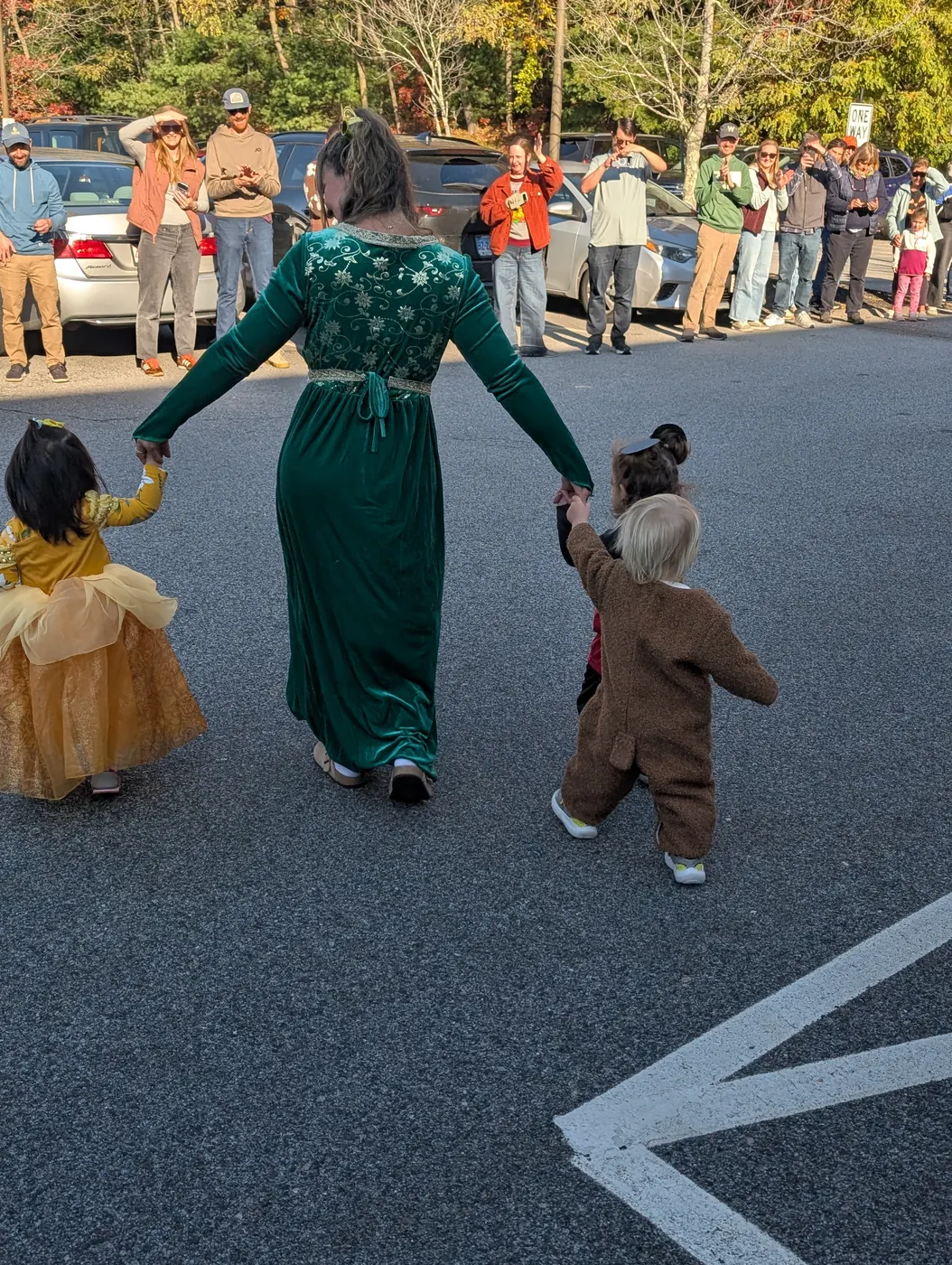 A grown-up in a green gown leading two toddlers in costume through the Halloween Parade