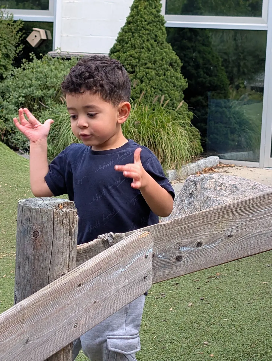 A preschooler talking with his hands by the wooden fence on the Blossom Station playground in Acton, MA