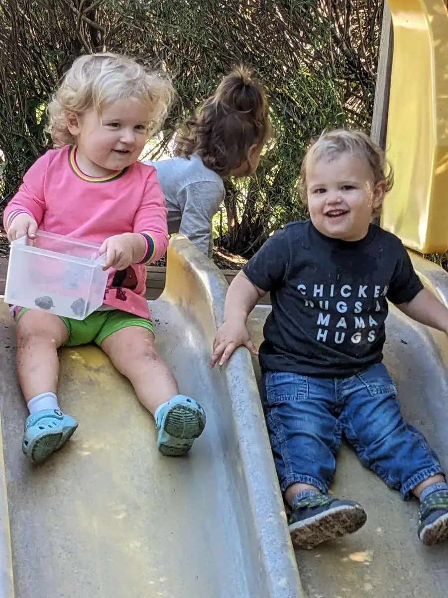 Two toddlers smiling together at the bottom of the slide at Blossom Station