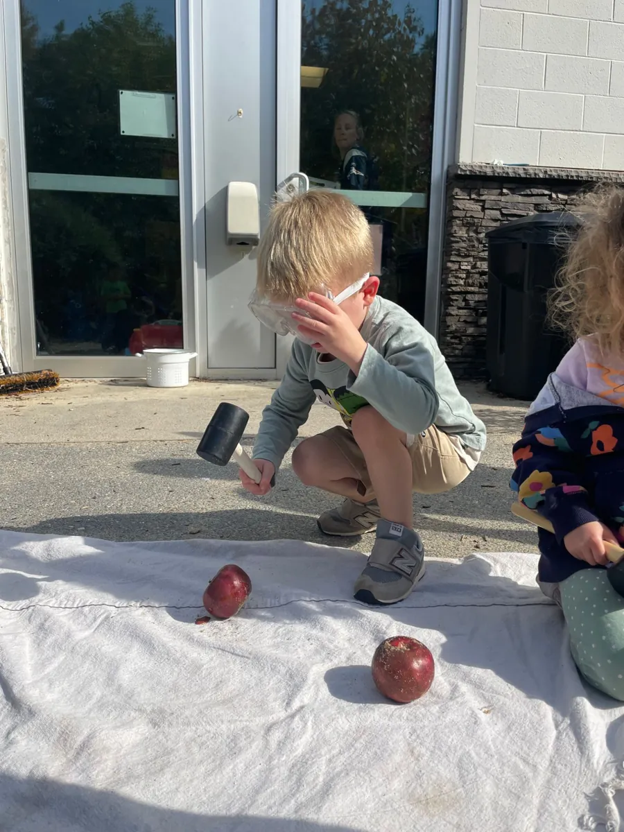 A preschooler in safety goggles crouching to tap an apple with a rubber mallet during TimberNook