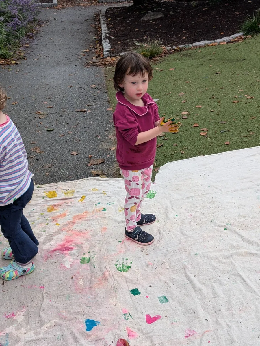 A toddler with paint-covered hands making prints on a big canvas outdoors during TimberNook at Blossom Station