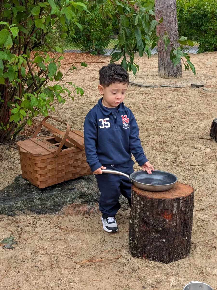 A toddler cooking with a frying pan on a tree stump in the TimberNook mud kitchen